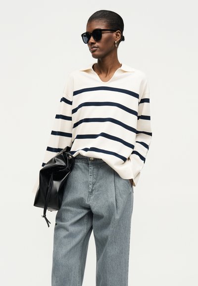 Γυναίκα που φοράει μαύρα γυαλιά ηλίου, κρεμ πουλόβερ με θαλασσί ρίγες, χαλαρό γκρι παντελόνι, κρατώντας μια μαύρη δερμάτινη τσάντα, στέκεται σε λευκό φόντο.