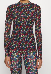 Frau in einem langärmeligen, durchsichtigen Oberteil und Leggings mit einem bunten kleinen Blumenmuster auf schwarzem Hintergrund.