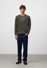 Man står mot en enfärgad vägg och bär en mörkgrön knappad cardigan, grå skjorta, blå jeans och bruna skor, med en hand i fickan.