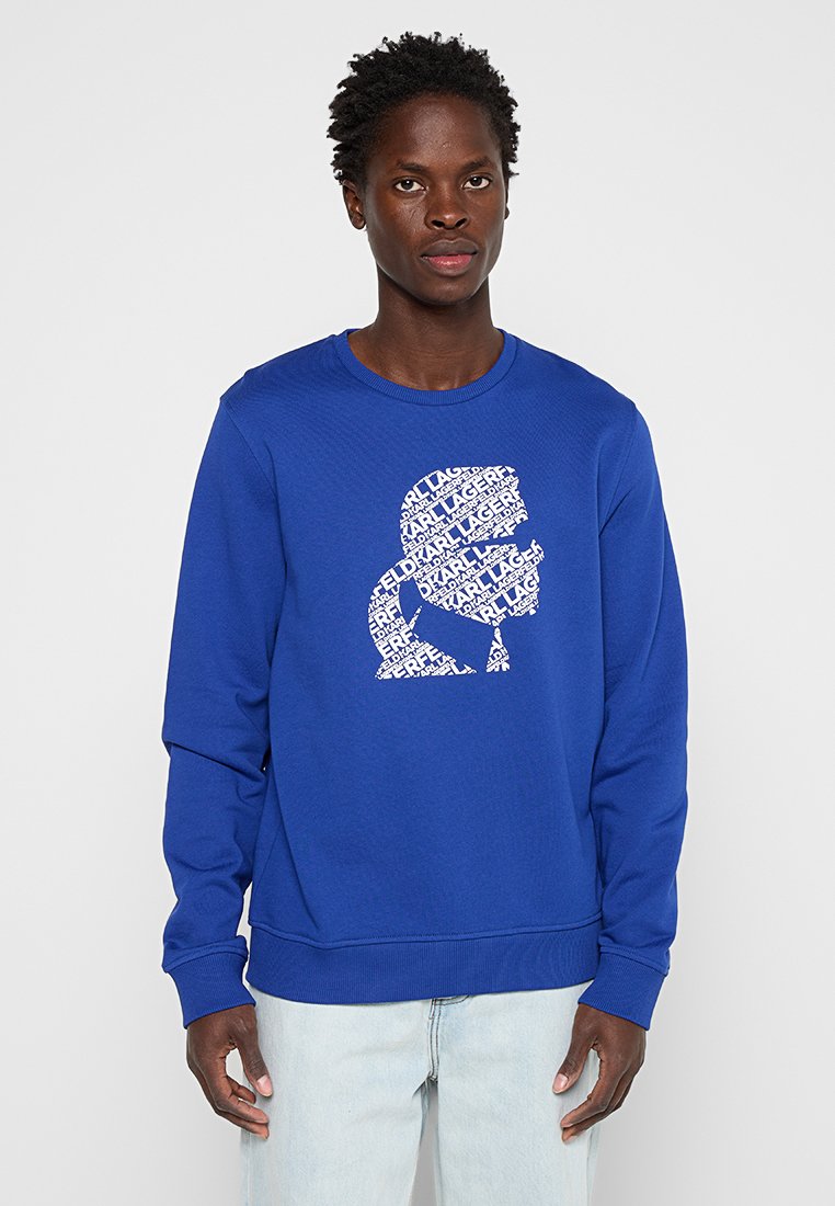 Un homme portant un sweat à capuche bleu avec un graphique blanc représentant un silhouette de tête composée de répétitions du texte "KARL LAGERFELD", associé à un jean clair.