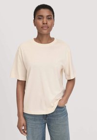 Beige Baumwoll-T-Shirt mit Rundhalsausschnitt und kurzen Ärmeln, mit einer lässigen Passform und glatter Textur. Getragen mit blauen Denim-Jeans.