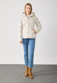 Doudoune beige clair avec capuche, présentant des coutures horizontales et un zip sur le devant. Associée à un jean bleu clair et des bottines en daim marron.