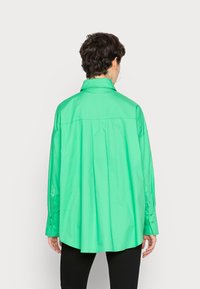 Camisa verde de botão, mangas longas, ajuste relaxado, tecido em mistura de algodão. Apresenta pregas nas costas, colarinho pontiagudo e detalhe costurado.