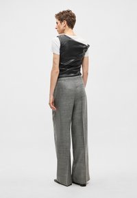 Colete de couro preto sobre uma t-shirt branca, combinado com calças cinzentas de padrão xadrez de cintura alta. Textura lisa, design estruturado, corte de perna larga.