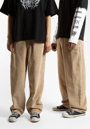 Pair of loose, beige corduroy pants with vertical ribbing, worn with black graphic t-shirts and black sneakers featuring white accents.