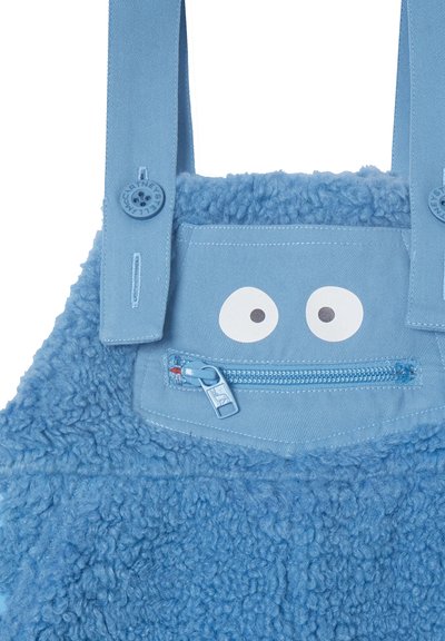 Salopette bleue pour enfants avec une texture duveteuse, bretelles ajustables avec boutons, une fermeture éclair sur la poche avant, et deux yeux de cartoon blancs au-dessus de la fermeture éclair.
