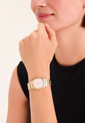 Montre en métal doré avec un cadran rectangulaire, un fond blanc, des index horaires dorés et un bracelet à maillons, portée sur un poignet.