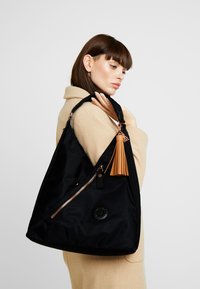 Black nylon shoulder bag with a matte finish, featuring a gold zipper, a leather tassel accent, and a circular logo patch.