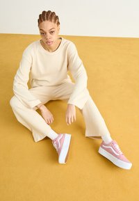 Rosa plattform sneakers med hvite detaljer, parret med en kremfarget, løstsittende langermet topp og matchende vide bukser, plassert på et gult gulv.
