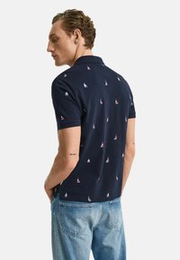 Polo shirt navy con piccoli motivi di barche a vela, maniche corte e colletto classico. Indossato con jeans in denim azzurro chiaro.