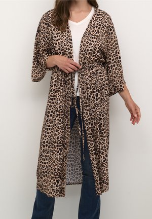 Femme portant un long cardigan style kimono à imprimé léopard noué à la taille sur un top blanc et un jean bleu, sur un fond uni.