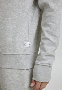 Sweat-shirt gris clair avec des poignets et un ourlet côtelés, arborant une étiquette latérale sur laquelle est inscrit "! SOLID". Le tissu est doux, avec une texture lisse.