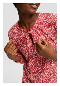 Blusa rossa a fiori con scollatura arricciata e chiusura con laccetto. Il tessuto è leggero, morbido al tatto e con maniche a tre quarti.