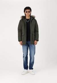 Olive green parka jacket with fur-lined hood, zip closure, two front pockets, worn over a black shirt and blue jeans, paired with white sneakers.