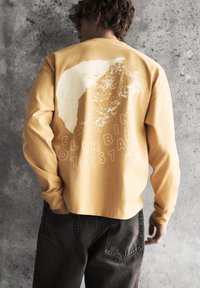 Personne aux cheveux bouclés portant un sweat-shirt moutarde avec un motif d'escalade et le texte "Grimper les étoiles" au dos.