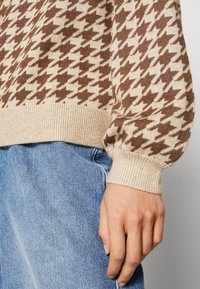 Stickad tröja i krämfärgad och brun hounds-tooth mönster. Har ribbade ärmslut och nederkant. Parat med ljusblå denimjeans.