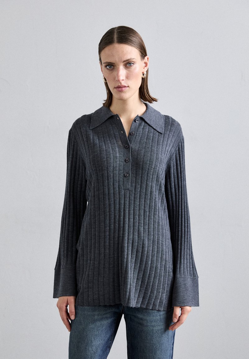 Camisola cinza, com ribas, de mangas longas e colarinho, apresentando uma frente com botões e punhos de mangas largas. Combinada com jeans de ganga azuis.