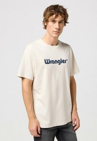 Beige-puuvillane t-särk, millel on suur tume sinine "Wrangler" logo rinnal. Lühikesed varrukad ja mugav lõige. Sile tekstuur.