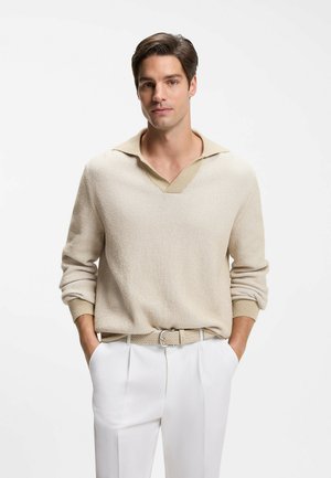 Ung mand iført beige striktrøje og hvide bukser med hænderne i lommerne, stående mod en ensfarvet hvid baggrund.