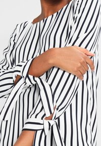 Camicia bianca con strisce verticali blu navy, dotata di scollatura a barca e maniche legate. Tessuto liscia con forma strutturata e senza motivi visibili.