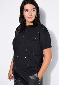 Camiseta negra de algodón, con adornos metálicos. Cuello redondo, mangas cortas y corte relajado. Llevada con pantalones de cuero negros.