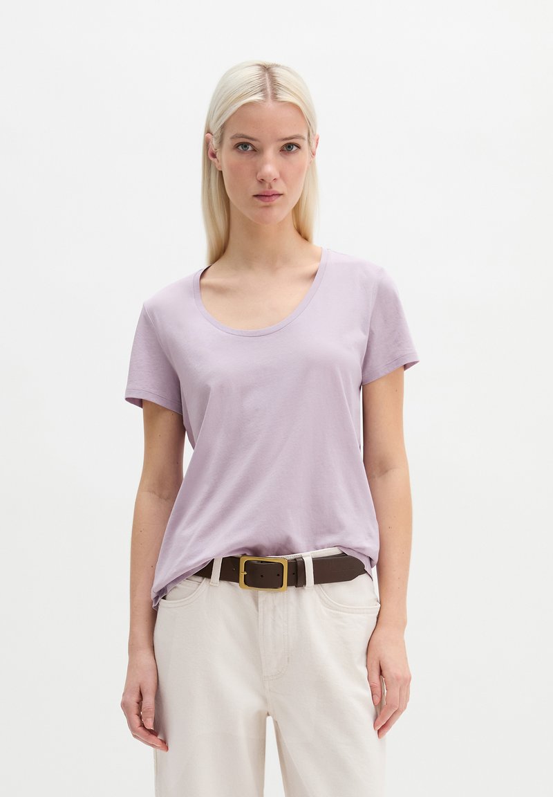 Junge Frau mit glattem blondem Haar, die ein hellviolettes kurzärmliges T-Shirt trägt, das in weiße Hosen mit braunem Gürtel gesteckt ist.