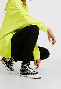 Felpa oversize di un luminoso verde lime con tasca, abbinata a pantaloni skinny neri e sneakers alte nere con testo bianco.