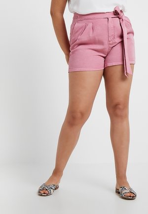 Femme portant un short rose taille haute avec poches latérales et ceinture nouée à la taille, associé à des sandales slip-on imprimé serpent.