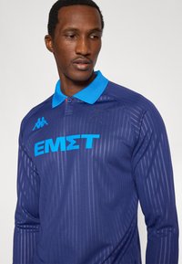 Polo a manica lunga blu navy con righe verticali, colletto blu e grande scritta blu "EMΣT". Presenta dettagli con il logo sul petto.