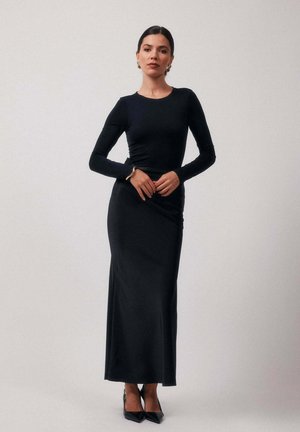 Robe noire à manches longues en tissu lisse, cintrée au niveau du corsage et s'évasant en une jupe ample. Des talons hauts noirs viennent parfaire ce look.