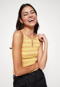 Top de alças amarelo com riscas, apresentando um detalhe com um buraco na gola, com alças finas e um design justo. Calças de ganga escuras com bordas desfiadas.