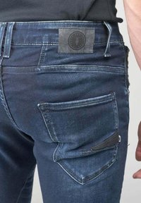 Los jeans de mezclilla azul oscuro presentan un parche en la cintura trasera, un bolsillo trasero con un acento negro y un detalle desgastado para una apariencia texturada.