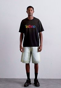 Tricou negru cu text „Techno” în culori curcubeu, pantaloni scurți cu un model deschis, șosete negre și pantofi închiși, purtat de un model masculin în picioare.