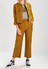 Senapsgul jacka med framfickor, kombinerad med en grå t-shirt med tryck och vida senapsgula byxor. Svarta och vita sneakers fulländar looken.