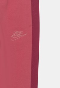 Tepláky Nike s růžovým hlavním tělem a bordó bočním panelem. Obsahují texturované detaily a kovové logo na přední straně.