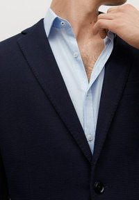 Granatowa marynarka o fakturze, z klapami, noszona na jasnoniebieskiej koszuli z kołnierzykiem, prezentująca zapięcie na dwa guziki oraz widoczne detale szycia.