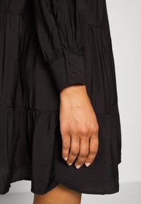 Svart lager-på-lager klänning med långa puffärmar. Har en slät textur och knappade manschetter, som visar upp distinkta lager av tyg.