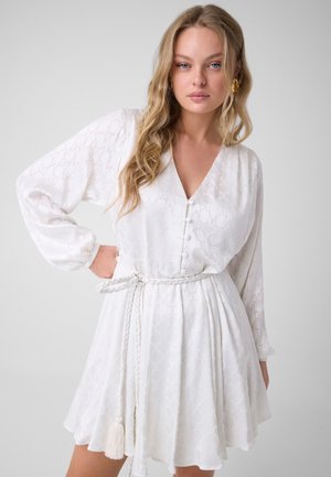 Vrouw met lang, golvend blond haar draagt een witte jurk met lange mouwen en een gevlochten ceintuur in de taille, staand tegen een effen lichte achtergrond.