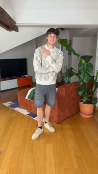 Ung mann smiler med armene i kors, iført en strikkegenser og shorts, står i en stue med sofa, TV og stor potteplante.