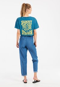 Mėlynos spalvos grafika t-shirt su užrašu "SAULĖTA POPIETĖ" geltonai, derinama su šviesiai mėlynomis džinsinėmis kelnėmis ir juodais šlepetėmis. Tekstūruota medžiaga, laisvas kirpimas.