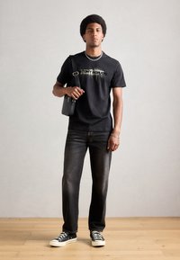 Tricou negru cu text grafic, purtat cu blugi închiși la culoare și teniși negri. Accesoriile includ o geantă neagră și un colier din lanț argintiu.