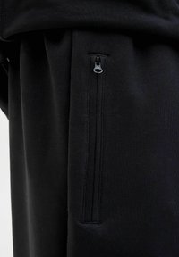 Pantaloni de trening negri cu un buzunar lateral cu fermoar, textura moale și o croială lejeră, concepuți pentru confort și practicitate.