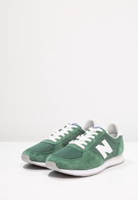 Gröna mockaskor med nätpaneler, vita snören och en framträdande vit "N"-logotyp på sidan. Gummisula med strukturerat grepp.