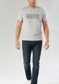 Camiseta gris de manga corta con texto negro en negrita "coronel tapiocca", combinada con jeans azul oscuro y zapatillas blancas. Aspecto sencillo y casual.