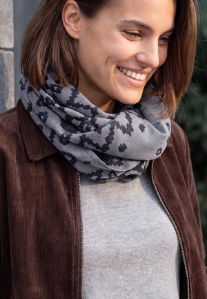 Une femme souriante en extérieur porte une écharpe grise à motif foncé, une veste marron et un haut gris.