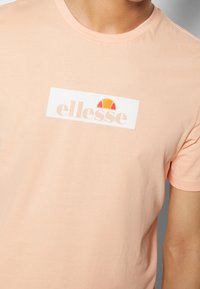 Meruňkový bavlněný tričko s bílým obdelníkovým logem a textem "ellesse" a polo-kru�žnicovou grafikou v oranžové a žluté barvě.