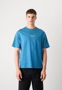 Kék pamut póló kerek nyakkal és rövid ujjakkal. A mellkason fehér "Calvin Klein Jeans" logó található. Lazán illeszkedő fazon.