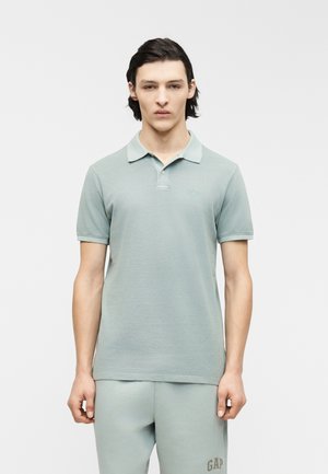 Jonge man die een lichtgroen poloshirt en bijpassende joggingbroek draagt, staand tegen een effen witte achtergrond.