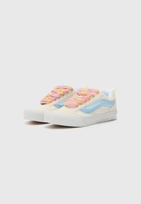 Low-top sneakers featuring a cream upper, blue side stripes, and multicolored woven laces. The sole is white with a textured surface.