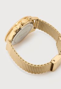 Ρολόι χειρός χρονογράφου σε χρυσό τόνο με υφασμένη υφή στο λουράκι και τρία πλευρικά κουμπιά, απεικονιζόμενο από πίσω που δείχνει το κούμπωμα και τις λεπτομέρειες της κάσας.